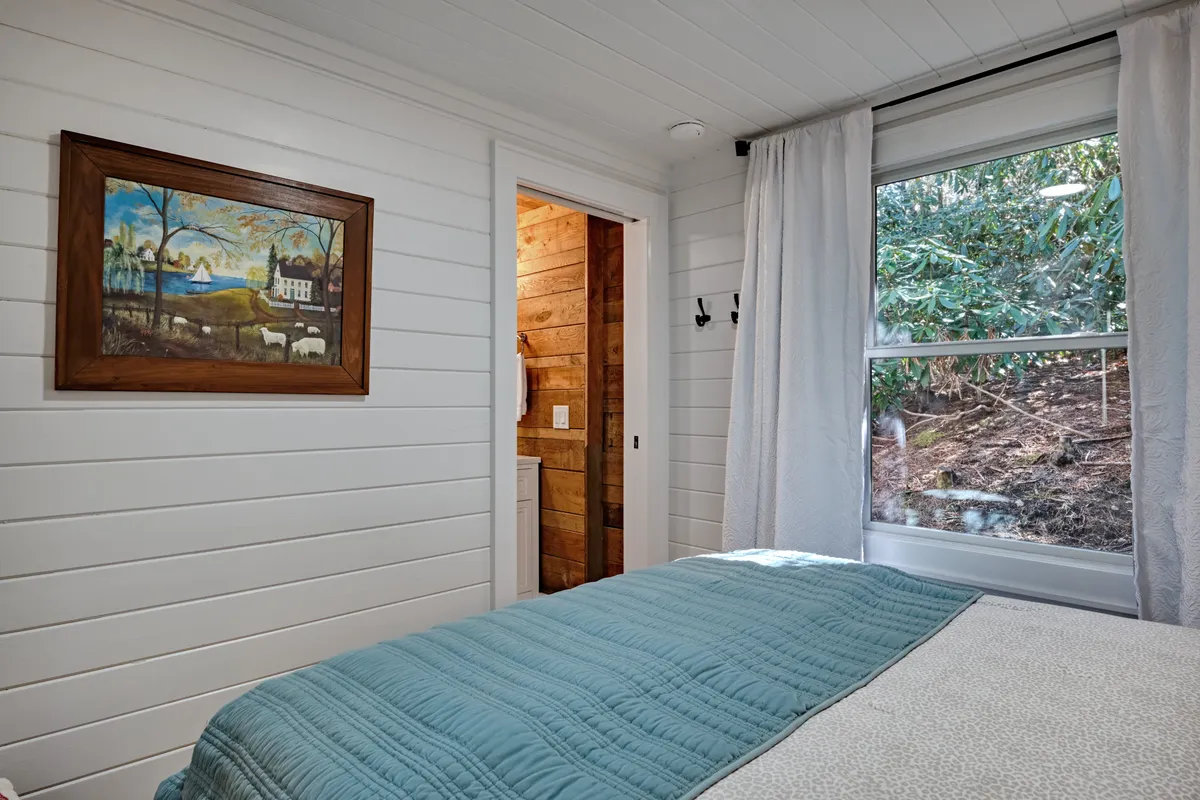 A peaceful bedroom with a bed dressed in a blue quilt, a painting on the wall, and soft natural light streaming in from a window. The space feels quiet and meditative, ideal for privacy and relaxation.