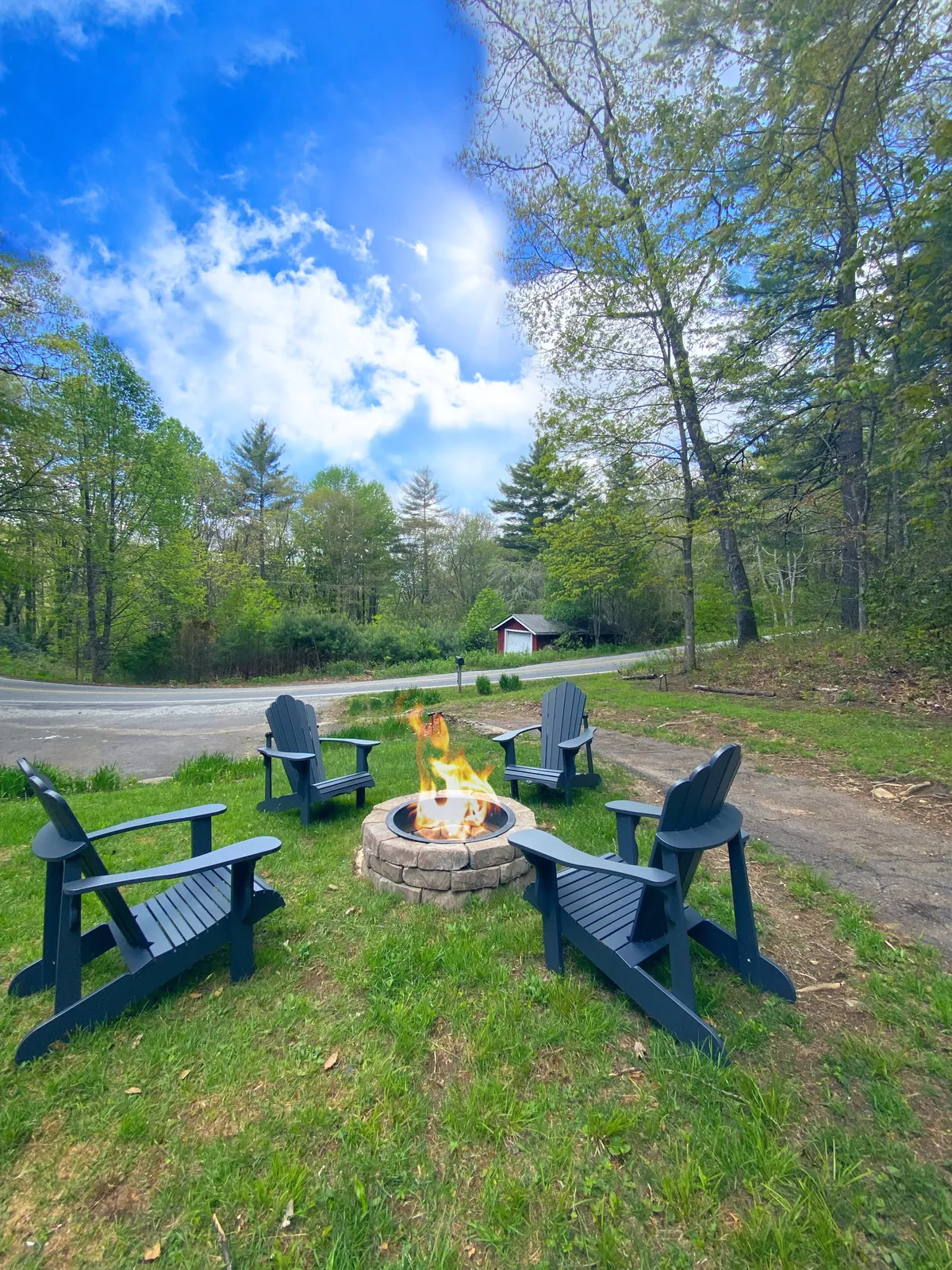 The hero image shows a lush green lawn bordered by tall trees under a vibrant blue sky. At the center is a newly built fire pit surrounded by cozy Adirondack chairs, inviting guests to gather, relax, and share stories as the sun sets or beneath a canopy of stars.