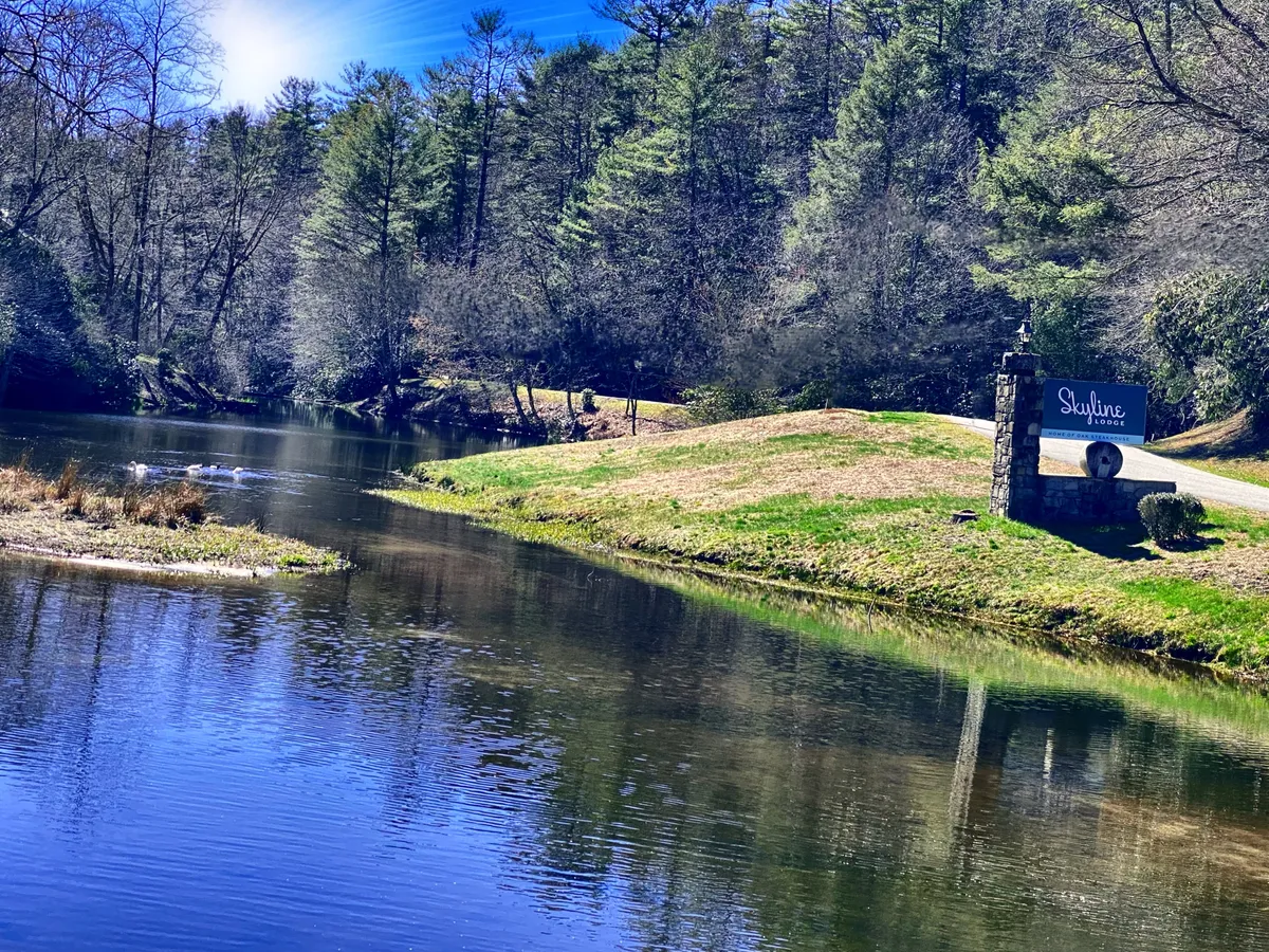 A serene exterior scene with a calm water surface, lush greenery, and a rustic stone signboard. The peaceful setting suggests opportunities for lakeside strolls, picnics, and enjoying nature just steps from the home.