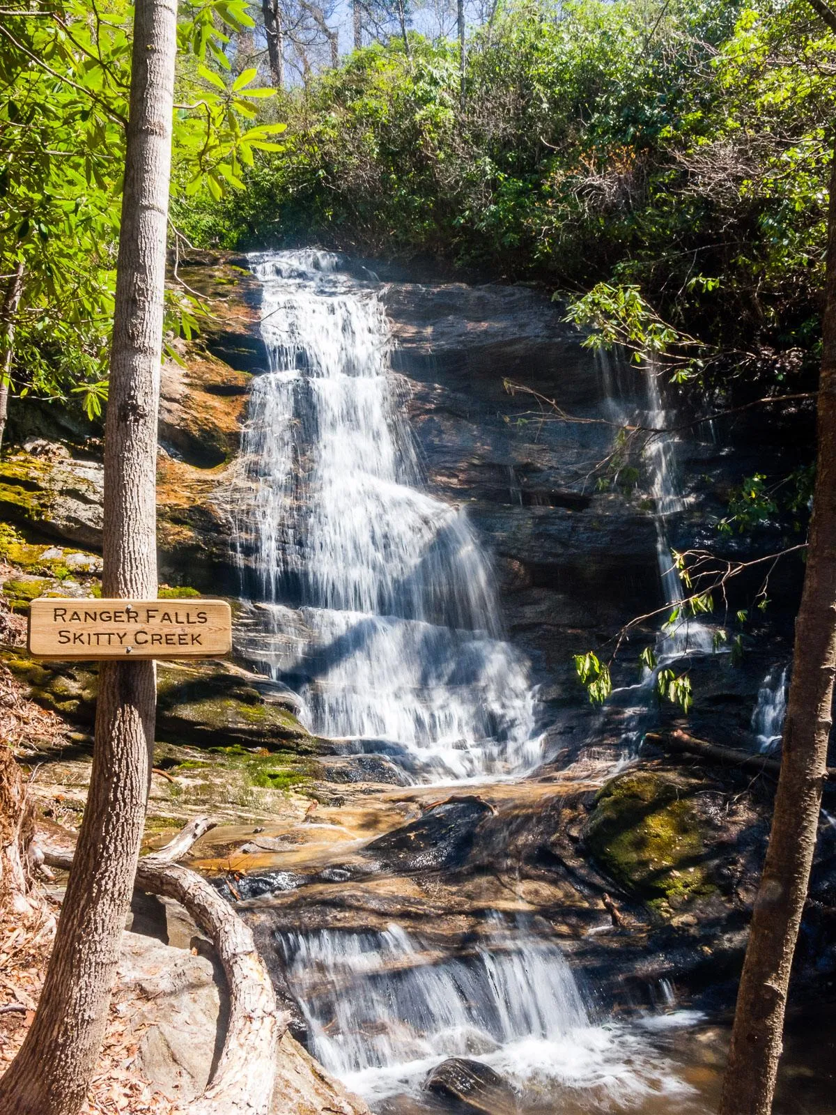 This image showcases a serene waterfall cascading over rock formations, surrounded by lush green foliage. A signpost at the site suggests easy access and marks the location as a point of interest for hikers and nature lovers. The tranquil mood and natural scenery emphasize the adventure and relaxation available nearby.