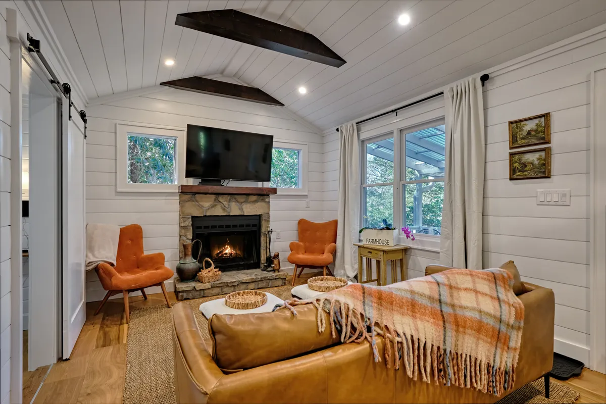 This image features a cozy living room with a fireplace (currently not in use), orange chairs, a sofa, and a flat-screen TV as focal points. The arrangement and decor create a warm, communal atmosphere perfect for relaxing, socializing, or movie nights. Generous natural light fills the space, and the open-plan layout is subtly evident from the spacious seating arrangement.