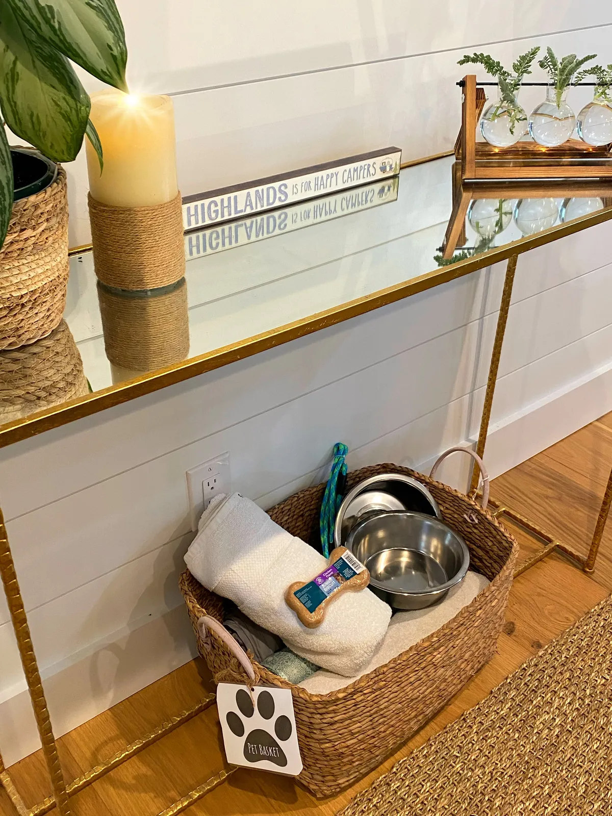 A warm, welcoming interior vignette featuring a golden framed mirror, a decorative candle, a lush green plant, and notably, a pet basket positioned near the entranceway. The arrangement suggests a pet-friendly environment, subtly indicating that four-legged guests are not just tolerated, but embraced in this space.