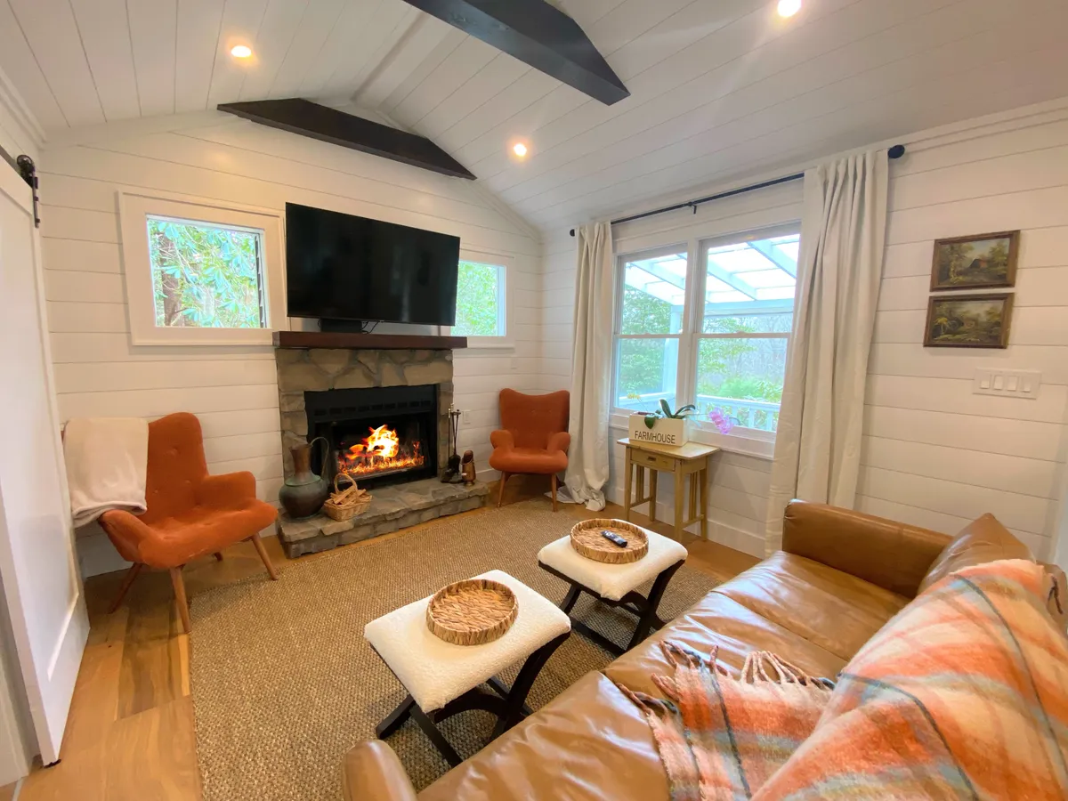 A warm and inviting interior living space featuring a fireplace, flat-screen television, armchairs, and a large window with a scenic view. The arrangement invites small groups to relax together after meals, enjoy a movie, or bask in the cozy ambiance.