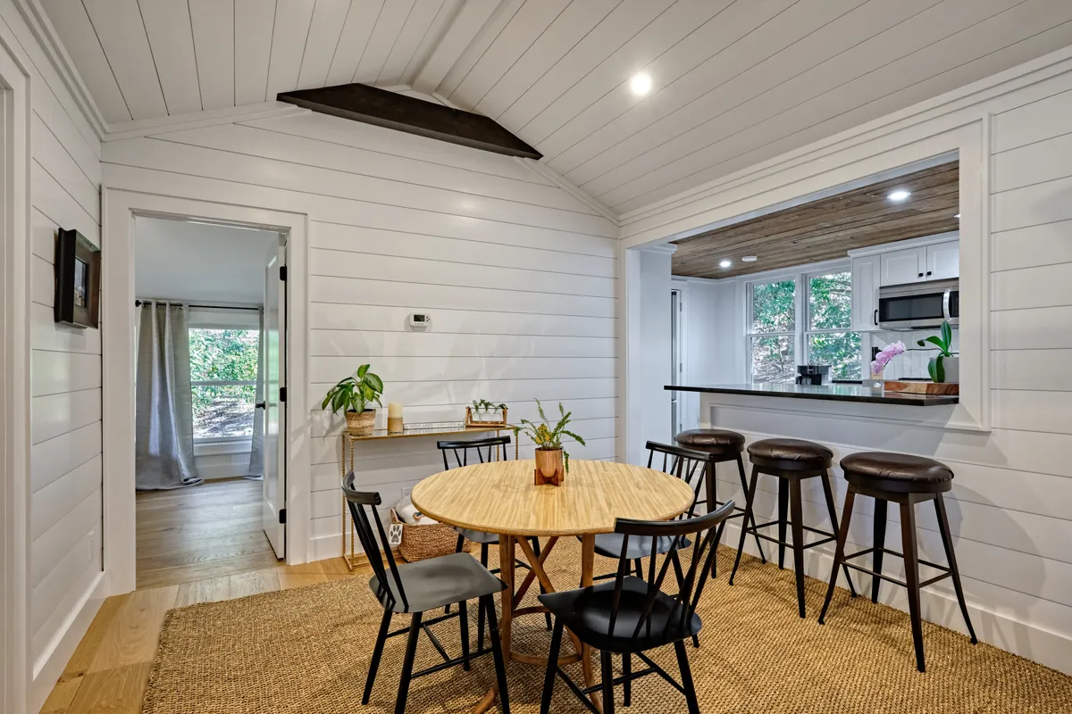 A round wooden dining table with black chairs sits next to an open kitchen area, bathed in natural light from surrounding windows. The scene conveys warmth and encourages gathering for meals, coffee chats, or group activities, blending seamlessly with the living space.