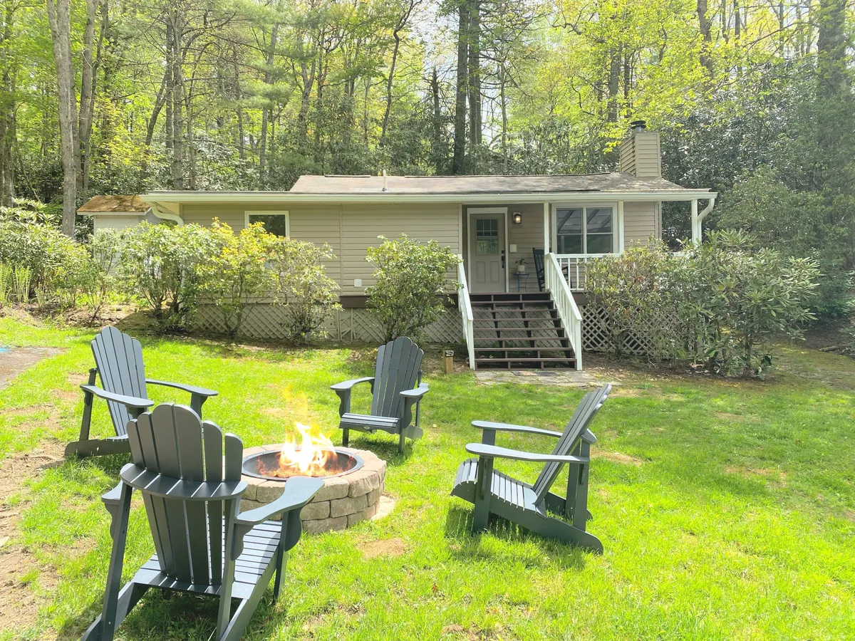 This image captures an inviting outdoor fire pit area surrounded by Adirondack chairs, with the front porch and lush trees in the background. The setting evokes scenes of gathering around the fire for warmth, relaxation, and conversation—perfect for fall and winter evenings. The porch is also visible, hinting at other ways to enjoy the outdoors.