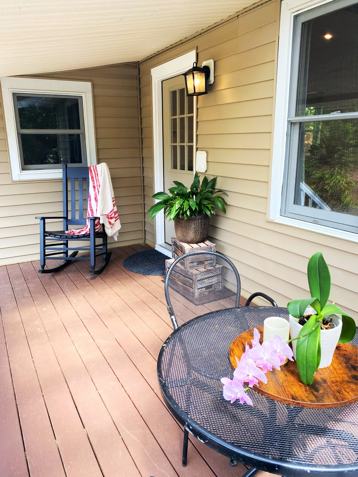 An inviting exterior scene featuring a rocking chair and a dining table set on a mountain-view patio, with potted plants and blooming orchid flowers adding to the welcoming atmosphere. The setup suggests a space ideal for gathering, relaxing with a book, or sharing morning coffee in the crisp mountain air.